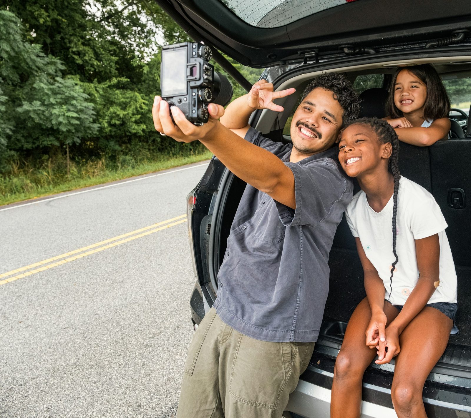 A father and his daughters taking a photo by their car on a road trip
