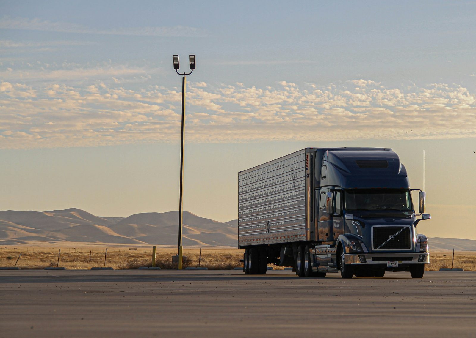 A dark blue semi truck parked in a desert lot at dusk, hills behind